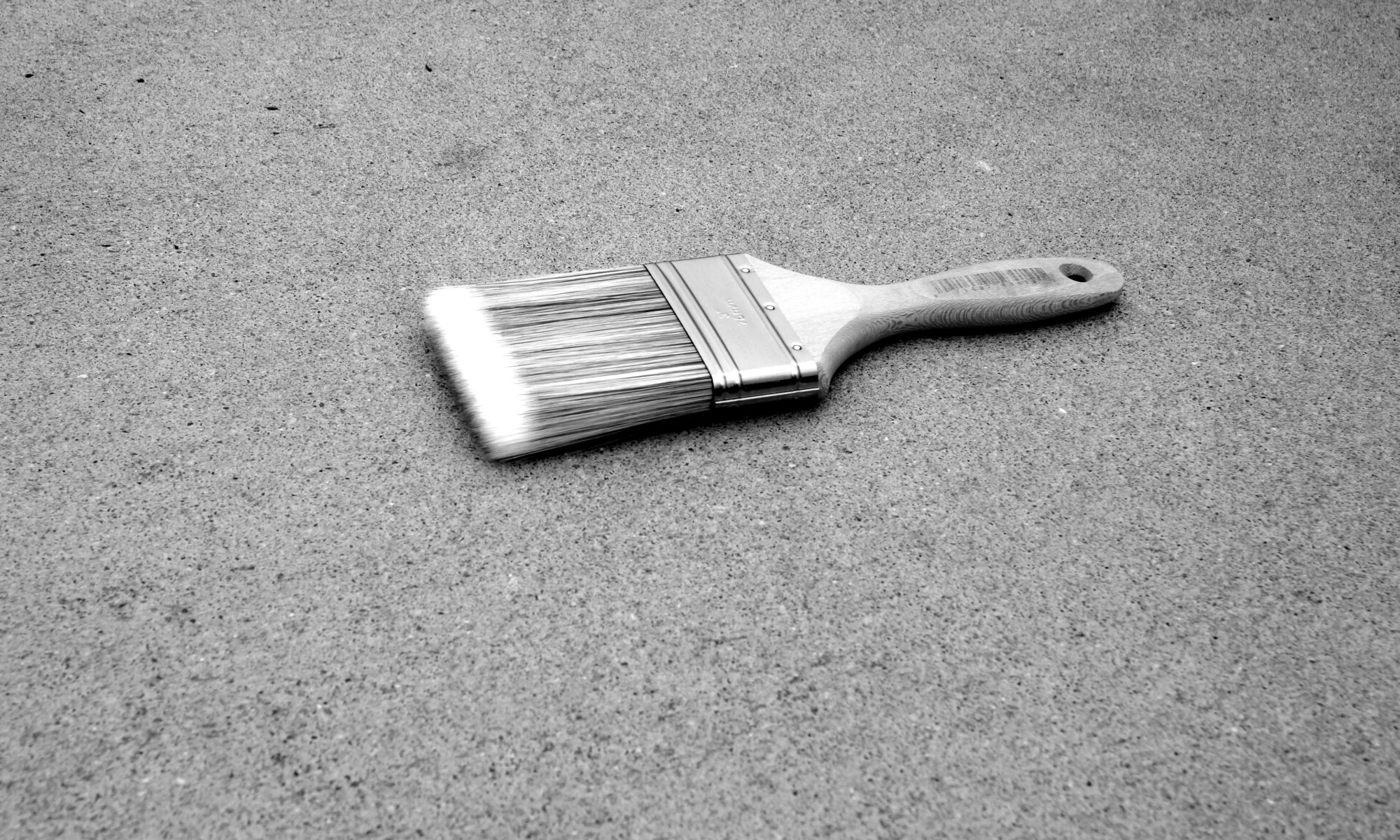 B&W oblique angle photo of a house-painting brush laying flat on a gritty concrete surface